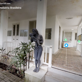 Imagem de um tour virtual no Memorial da Pediatria Brasileira. Em primeiro plano, há uma estátua em tom escuro representando uma mulher em pé segurando um bebê nos braços. A estátua está posicionada ao lado de uma pequena escada que leva a uma varanda coberta. A varanda possui piso decorado, colunas brancas e portas e janelas também brancas. Placas comemorativas estão fixadas na parede ao fundo. À direita, um corredor aberto segue ao longo da fachada, onde se vêem painéis expositivos afixados na parede. Plantas ornamentais ocupam a área ao lado da escada. O ambiente transmite a aparência de um espaço histórico e institucional.
