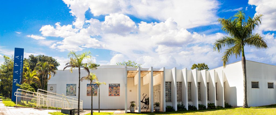 Fachada da frente do Museu em dia ensolarado, com destaque para duas palmeiras ao redor do prédio, que se caracteriza por sua estrutura moderno, nas cores branco e azul.