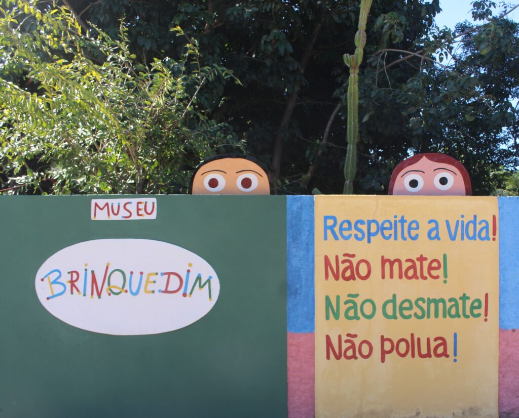 o Descrição Geral: A imagem mostra um muro colorido, cercado por uma vegetação densa de árvores verdes e um cacto alto ao fundo, sob a luz do sol. O muro é dividido em seções de cores vibrantes e por cima se vê cabeças de bonecos que olham para o lado de fora. O da esquerda tem cabelos pretos e o da direita cabelos avermelhados. Ambos possuem olhos brancos muito grandes e redondos, com pupilas pretas, criando uma expressão de curiosidade e surpresa. Detalhes da Composição: Do lado Esquerdo a parede é pintada de verde escuro. No topo, há uma pequena placa retangular branca com a palavra "MUSEU" em letras vermelhas. Logo abaixo, há um grande oval branco que contém o nome "BRINQUEDIM". As letras são estilizadas, coloridas (azul, amarelo, vermelho e verde) e possuem pequenos pontos nas extremidades. Do lado Direito tem um Mural de Mensagens o Esta seção tem um fundo amarelo claro, com bordas laterais em azul e uma base rosa. o Nela, está escrita a seguinte mensagem em letras grandes, cada linha em uma cor diferente: Respeite a vida! (em azul) Não mate! (em vermelho) Não desmate! (em verde) Não polua! (em marrom/bordô)