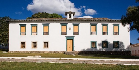 Fotografia da fachada frontal do museu, um edifício de dois andares, com paredes brancas, janelas retangulares com moldura verde e venezianas amarelas. No centro, há uma porta de madeira com moldura verde e, acima dela, um pequeno brasão na parede. No topo do telhado, há uma pequena mansarda. Em frente ao edifício, há uma faixa de grama e calçamento. O céu está azul com algumas nuvens.
