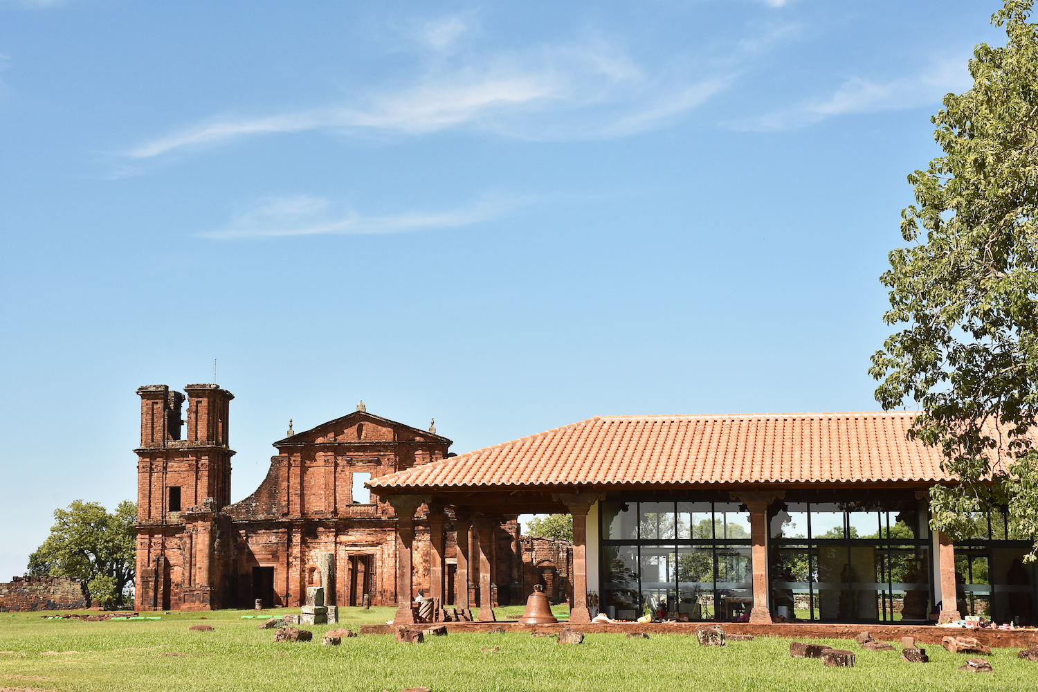 Fotografia do museu, com as ruínas da Igreja São Miguel Arcanjo ao fundo. A ruína exibe uma torre lateral e paredes de pedra em tons avermelhados, ressaltando seu caráter majestoso. Em primeiro plano, o Museu, um edifício contemporâneo com cobertura de telhas cerâmicas e paredes de vidro, avarandado em todo seu perímetro com destaque para pilares em pedra, apoiando a cobertura.