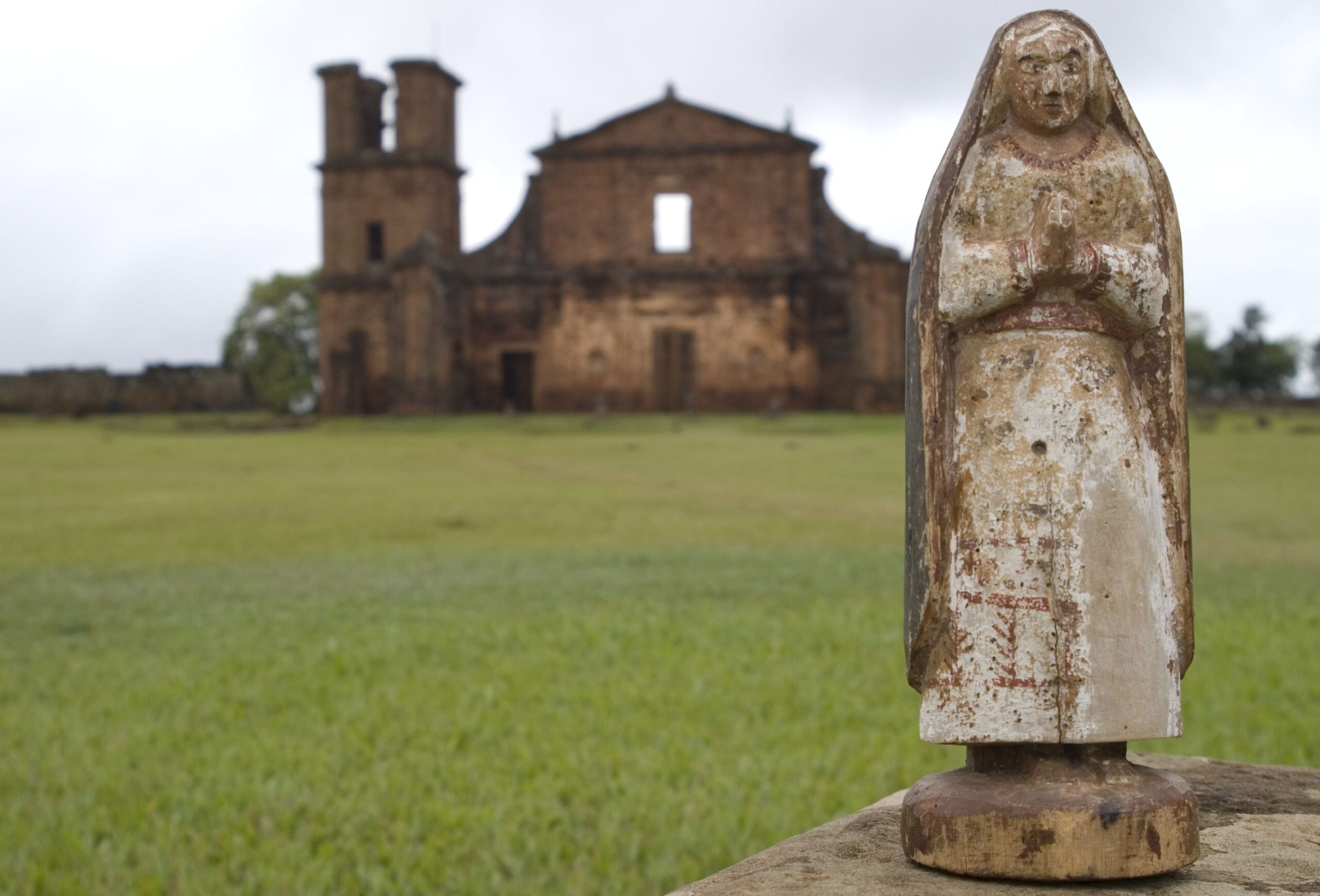 Fotografia da área gramada do Museu, apresentando uma estátua de figura religiosa em destaque, posicionada em primeiro plano. Ao fundo, de forma desfocada, é possível observar as ruínas da Igreja São Miguel Arcanjo, compondo um cenário que mistura elementos históricos e religiosos.