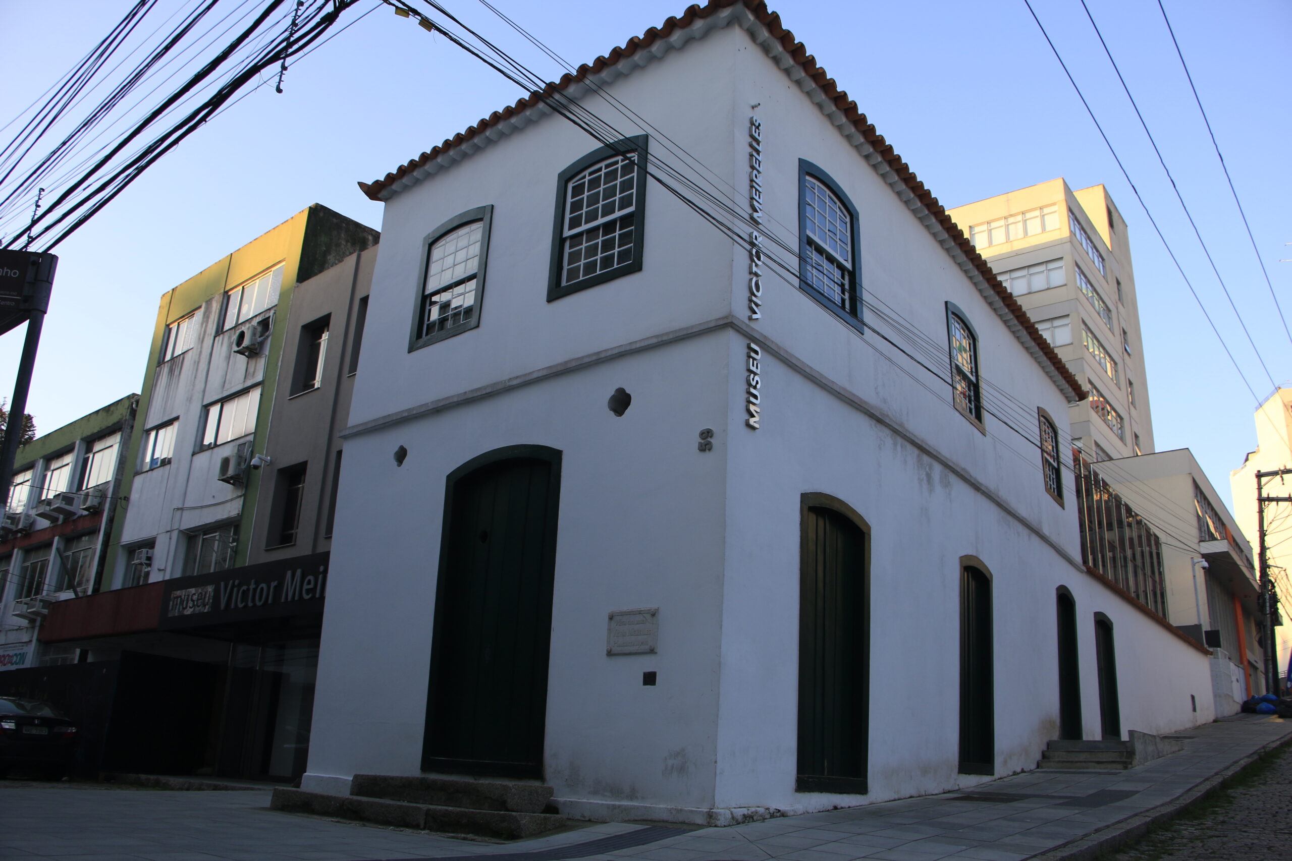Fotografia da fachada do Museu Victor Meirelles, edificação de dois pavimentos, pintada em branco, com janelas tipo guilhotina em madeira e vidro, emolduradas por perfis pintados em verde. A cobertura é aparente e em telhas cerâmicas. Há uma placa vertical na fachada que exibe o nome 'Museu Victor Meirelles'. Ao fundo, há outros prédios e fios elétricos cruzando o céu.