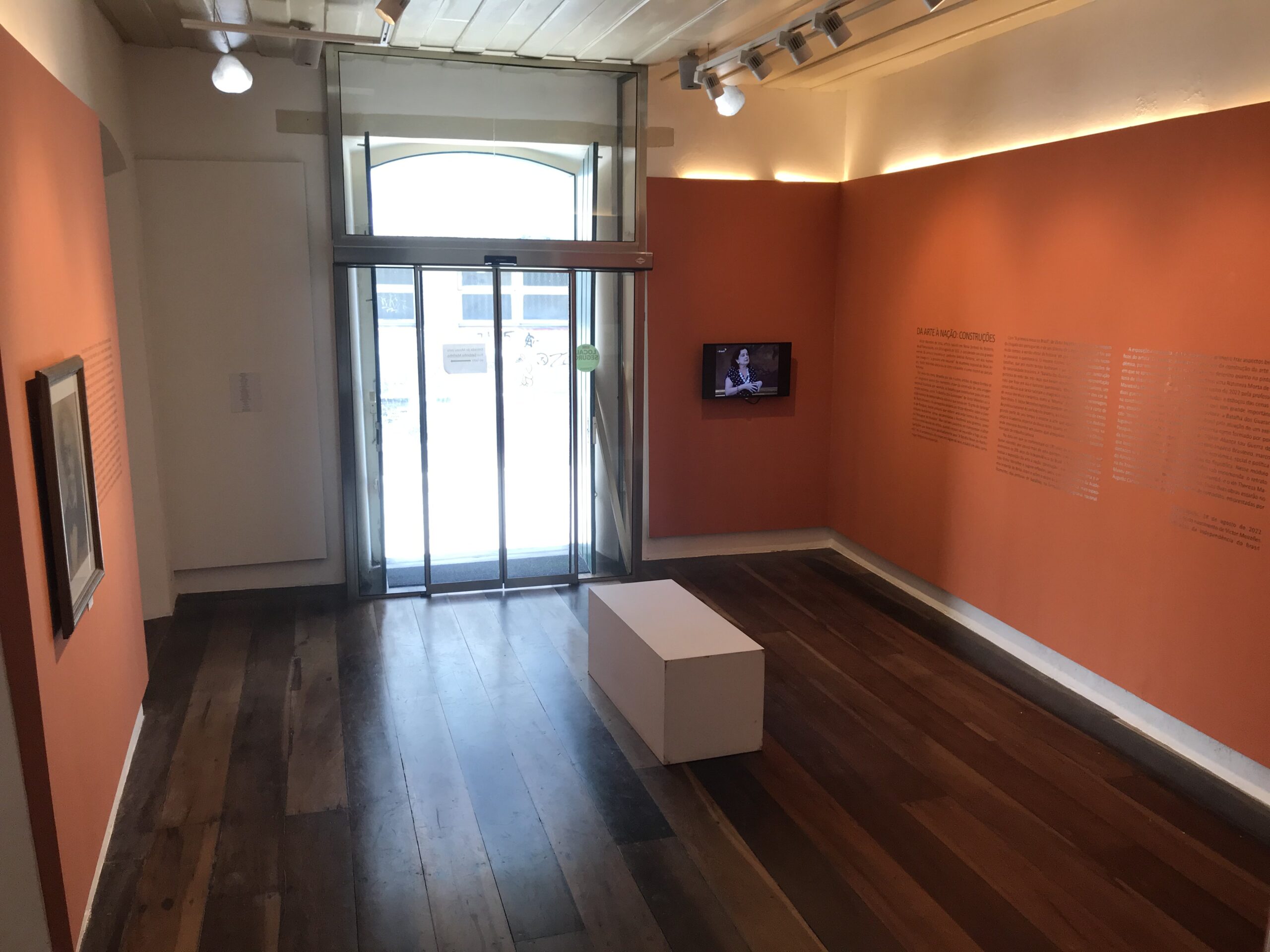 Fotografia de espaço expositivo com paredes pintadas na cor laranja e piso de madeira. À esquerda, observa-se uma obra de arte emoldurada e fixada na parede. No centro da sala, há um banco branco de formato retangular. Na parede do fundo, à direita de uma janela, encontra-se uma televisão que exibe um vídeo, acompanhada por um texto explicativo fixado na parede ao lado.