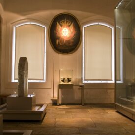 Sala de exposição com paredes e pisos claros. Na lateral direita, nota-se vitrine de vidro de grande altura, com vários objetos expostos; na esquerda, duas lápides em pedra com escritos. Ao fundo, duas esquadrias em arco abatido estão fechadas com persianas e entre elas, no alto, um brasão de armas de formato oval iluminado e, embaixo, uma vitrine com objeto.