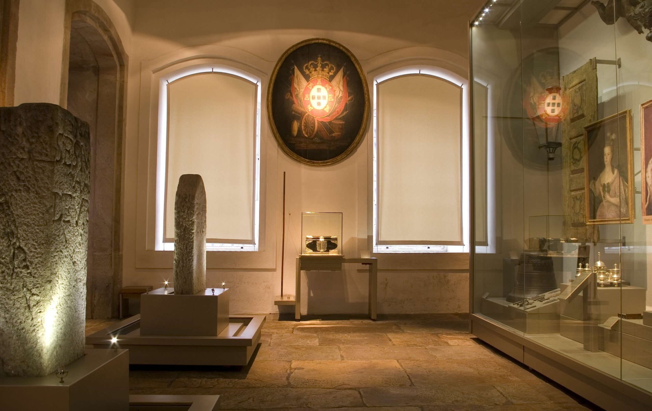 Sala de exposição com paredes e pisos claros. Na lateral direita, nota-se vitrine de vidro de grande altura, com vários objetos expostos; na esquerda, duas lápides em pedra com escritos. Ao fundo, duas esquadrias em arco abatido estão fechadas com persianas e entre elas, no alto, um brasão de armas de formato oval iluminado e, embaixo, uma vitrine com objeto.