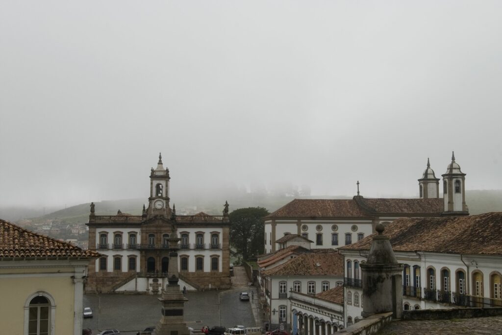 Fotografia com vista do Museu e do seu entorno imediato, com construções coloniais, destacando suas fachadas, janelas em arcos e telhados cerâmicos, com uma mescla de cores que estabelece um padrão visual de cidade histórica. A fachada do Museu da Inconfidência é monumental e imponente, com volume em quatro níveis. Há simetria na composição da fachada com entrada demarcada ao centro sob escadaria e nas laterais as janelas e balcões, mostrando o ritmo de um projeto com uma estética clássica e de decoração elaborada. Ao topo, uma torre com sino se destaca no centro da fachada.
