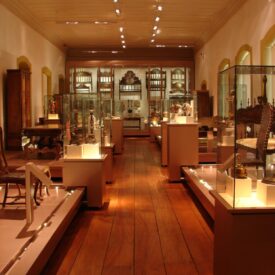 Sala de exposição, de forma retangular, comprida e de grande vão, com teto e parede clara e piso em madeira. Possui um corredor central ladeado por vitrines de diferentes formatos que contêm uma variedade de objetos, em termos de forma, material e tamanho. A iluminação acompanha o layout do mobiliário com luminárias direcionadas aos objetos.