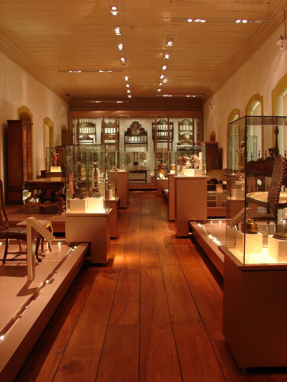 Sala de exposição, de forma retangular, comprida e de grande vão, com teto e parede clara e piso em madeira. Possui um corredor central ladeado por vitrines de diferentes formatos que contêm uma variedade de objetos, em termos de forma, material e tamanho. A iluminação acompanha o layout do mobiliário com luminárias direcionadas aos objetos.