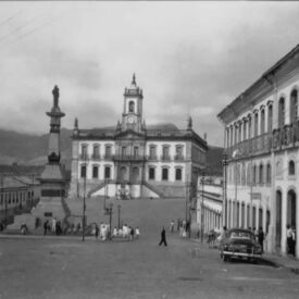 Fotografia histórica, em preto e branco, da Praça Tiradentes, praça central com obelisco, circundada por ruas em paralelepípedos, com vários edifícios históricos ao redor e ao fundo o Museu da Inconfidência. Os edifícios são, na sua maioria, de dois pavimentos, com estilo de arquitetura colonial.