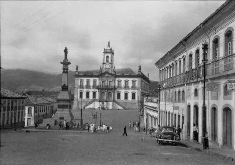 Fotografia histórica, em preto e branco, da Praça Tiradentes, praça central com obelisco, circundada por ruas em paralelepípedos, com vários edifícios históricos ao redor e ao fundo o Museu da Inconfidência. Os edifícios são, na sua maioria, de dois pavimentos, com estilo de arquitetura colonial.