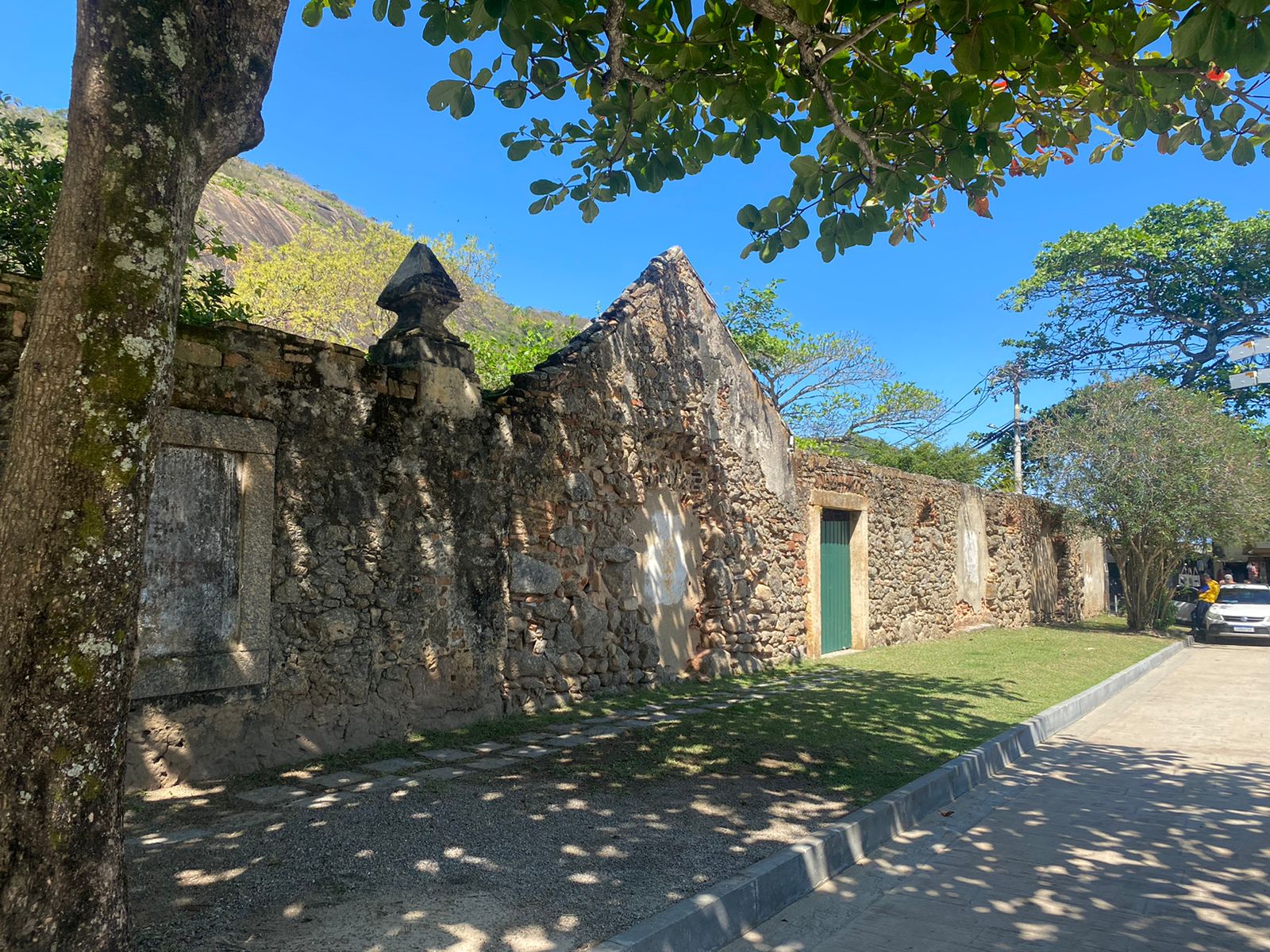 Fotografia da fachada do museu, composta por extenso muro de pedras com aberturas ao longo dele, com destaque para um portão de madeira com pintura verde, que é o acesso do Museu. A fachada apresenta um frontão em pedra e pequenos elementos decorativos. Em frente, há uma via de paralelepípedos e uma calçada revestida com grama, além da presença de árvores de grande porte criando sombras sobre a estrutura de pedra.