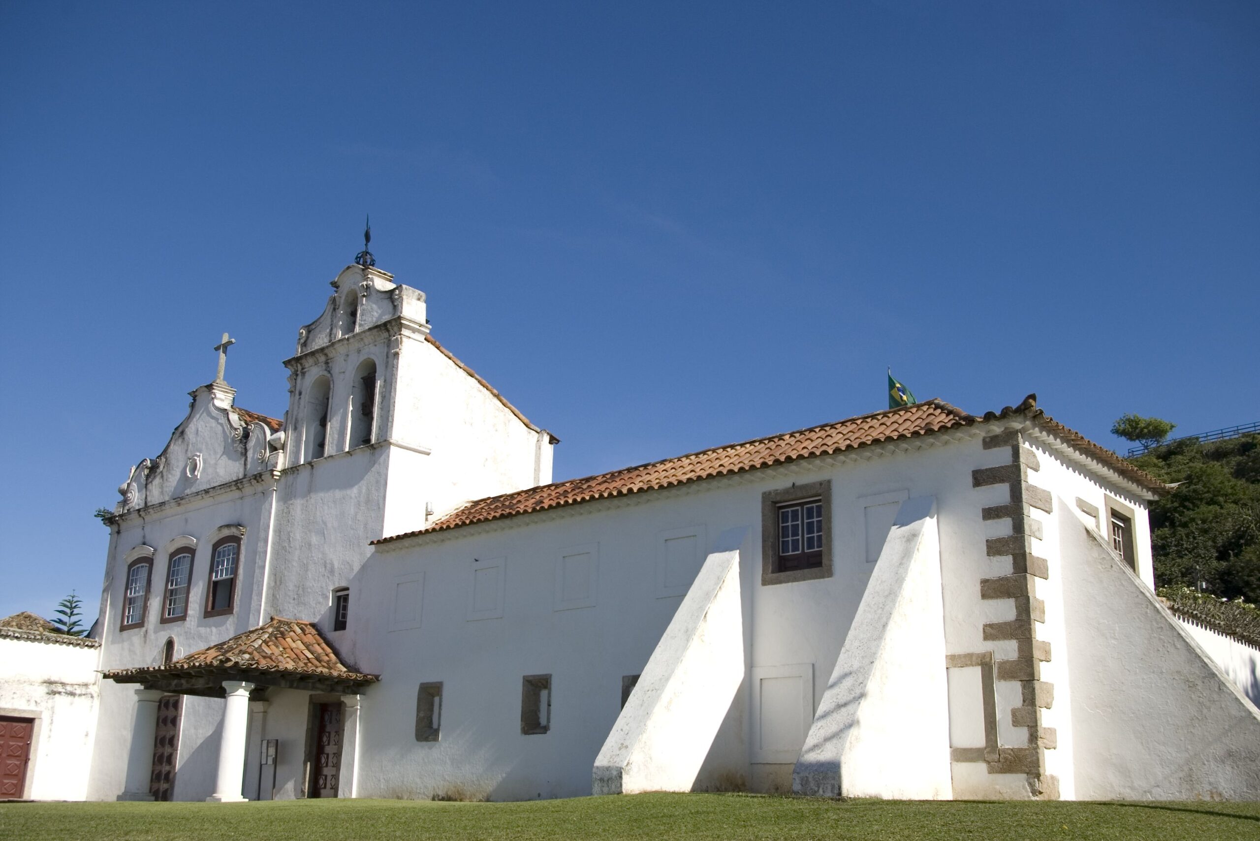 Fotografia do museu, destacando sua fachada branca com cobertura em telhas cerâmicas. Na parte esquerda da imagem está a Igreja, construção que possui uma torre sineira que se destaca em altura dos demais volumes. No térreo da Igreja, a entrada é emoldurada por alpendre sustentado por colunas simples. Na parte direita a ala frontal é composta por volume retangular de dois pavimentos e cobertura cerâmica em quatro águas.