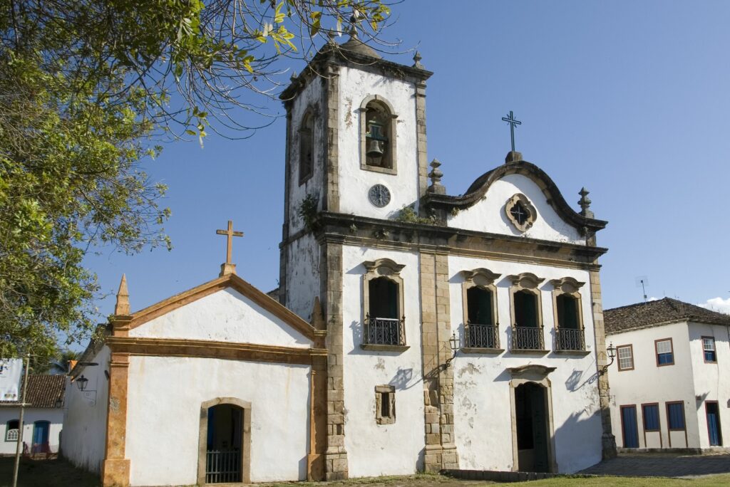 Fotografia da fachada frontal da Igreja de Santa Rita. A construção apresenta paredes brancas com detalhes nas extremidades em pedra, uma torre sineira à esquerda e um frontão ornamentado no corpo central. À esquerda da igreja, há uma fachada simples com frontão triangular e cruz no topo. O céu está azul e galhos de árvore estão parcialmente visíveis no canto superior esquerdo. Ao fundo, vê-se parte das construções históricas do centro da cidade.