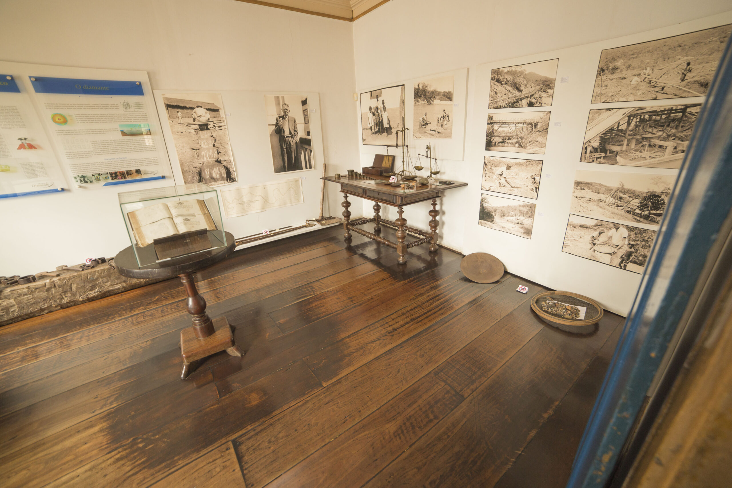 Fotografia de sala expositiva com vários quadros e fotografias históricas expostos nas paredes. Alguns itens estão expostos em mobiliários antigos. As paredes são brancas, em contraste com os pisos e móveis de madeira escura.