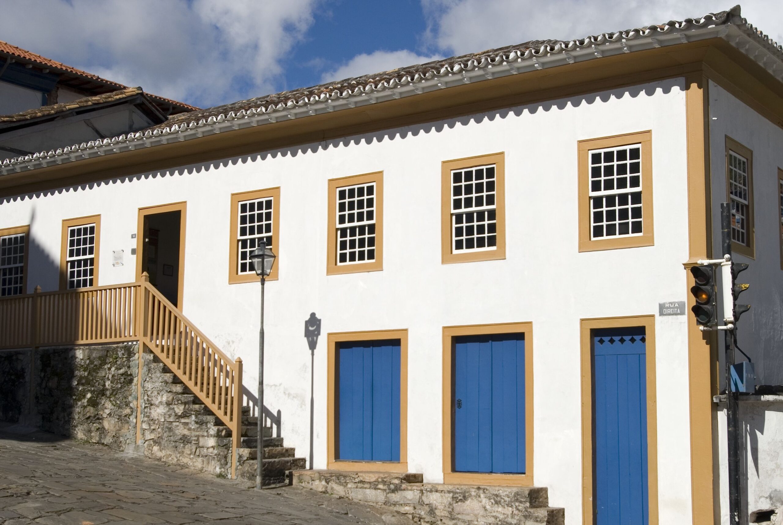 Fotografia da fachada do Museu, edifício colonial com pintura branca. O edifício possui uma série de janelas e portas em um padrão simétrico. As portas, no térreo, são azuis com molduras em amarelo, ladeadas por escada de pedra com guarda-corpo amarelo. As janelas tipo guilhotina, localizadas no primeiro pavimento, possuem molduras amarelas, contrastando com o branco da fachada.