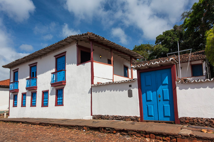 A imagem apresenta o casarão em perspectiva, com detalhe para a fachada principal e lateral direita do prédio. A edificação possui dois andares, de paredes brancas e molduras avermelhadas com esquadrias pintadas em azul. No térreo, há cinco janelas distribuídas de modo simétrico e, no pavimento superior, três portas avarandadas são ornadas por balcões em balaústre. Ao fundo, notam-se árvores sob um céu parcialmente nublado.
