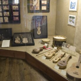 Fotografia de vitrine de grande dimensão com revestimentos claros. Nela estão expostos vários instrumentos musicais, incluindo violino, flauta, tambor e violão. Os instrumentos são de madeira e mostram sinais de uso. Há também alguns instrumentos musicais e ferramentas usadas para a fabricação de instrumentos. No fundo da vitrine estão dispostos quadros com fotografias.