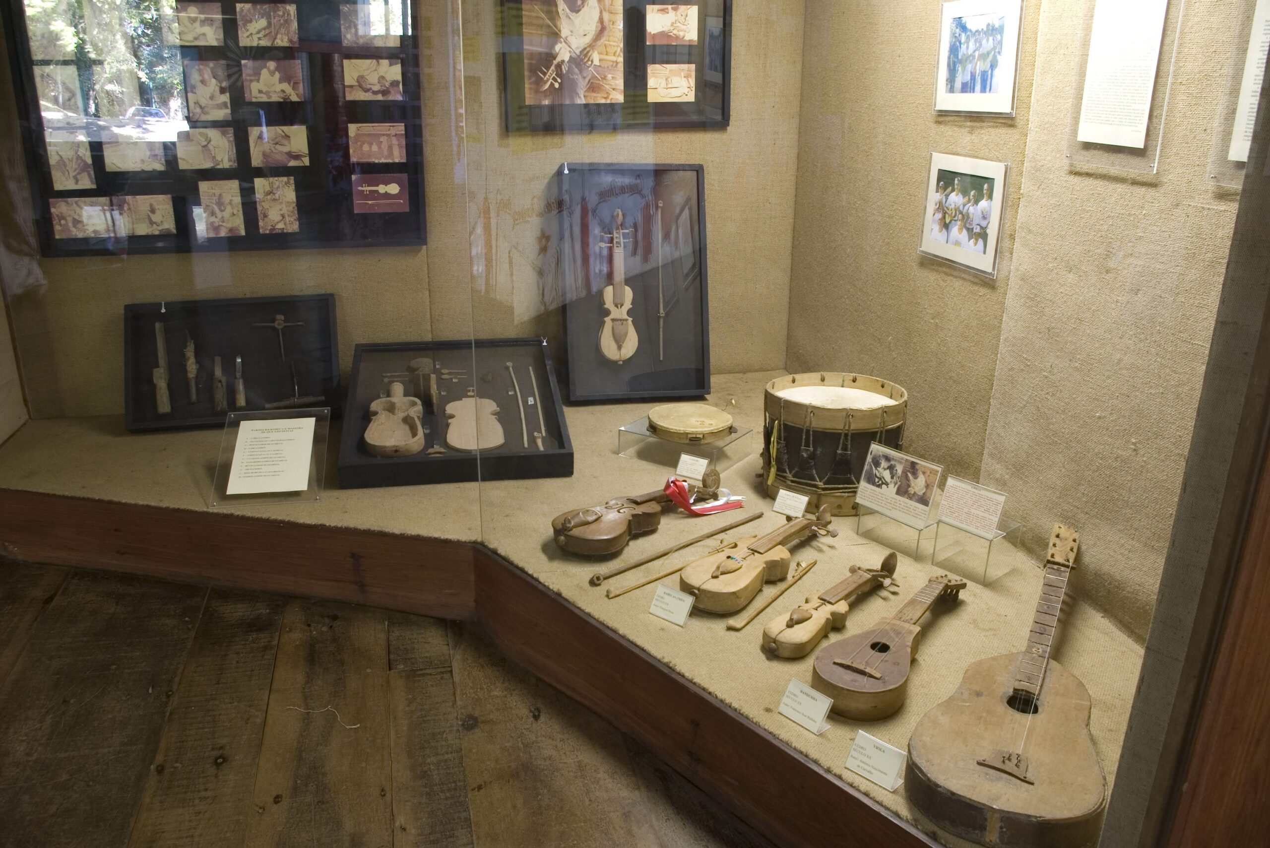 Fotografia de vitrine de grande dimensão com revestimentos claros. Nela estão expostos vários instrumentos musicais, incluindo violino, flauta, tambor e violão. Os instrumentos são de madeira e mostram sinais de uso. Há também alguns instrumentos musicais e ferramentas usadas para a fabricação de instrumentos. No fundo da vitrine estão dispostos quadros com fotografias.