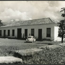 Fotografia histórica em preto e branco, retratando o prédio do museu. No primeiro plano, há um grande gramado e, em frente ao edifício, um carro, um fusca, modelo clássico automobilístico. Ao fundo, grandes nuvens tapam a vista dos morros, caracterizando o dia nublado.