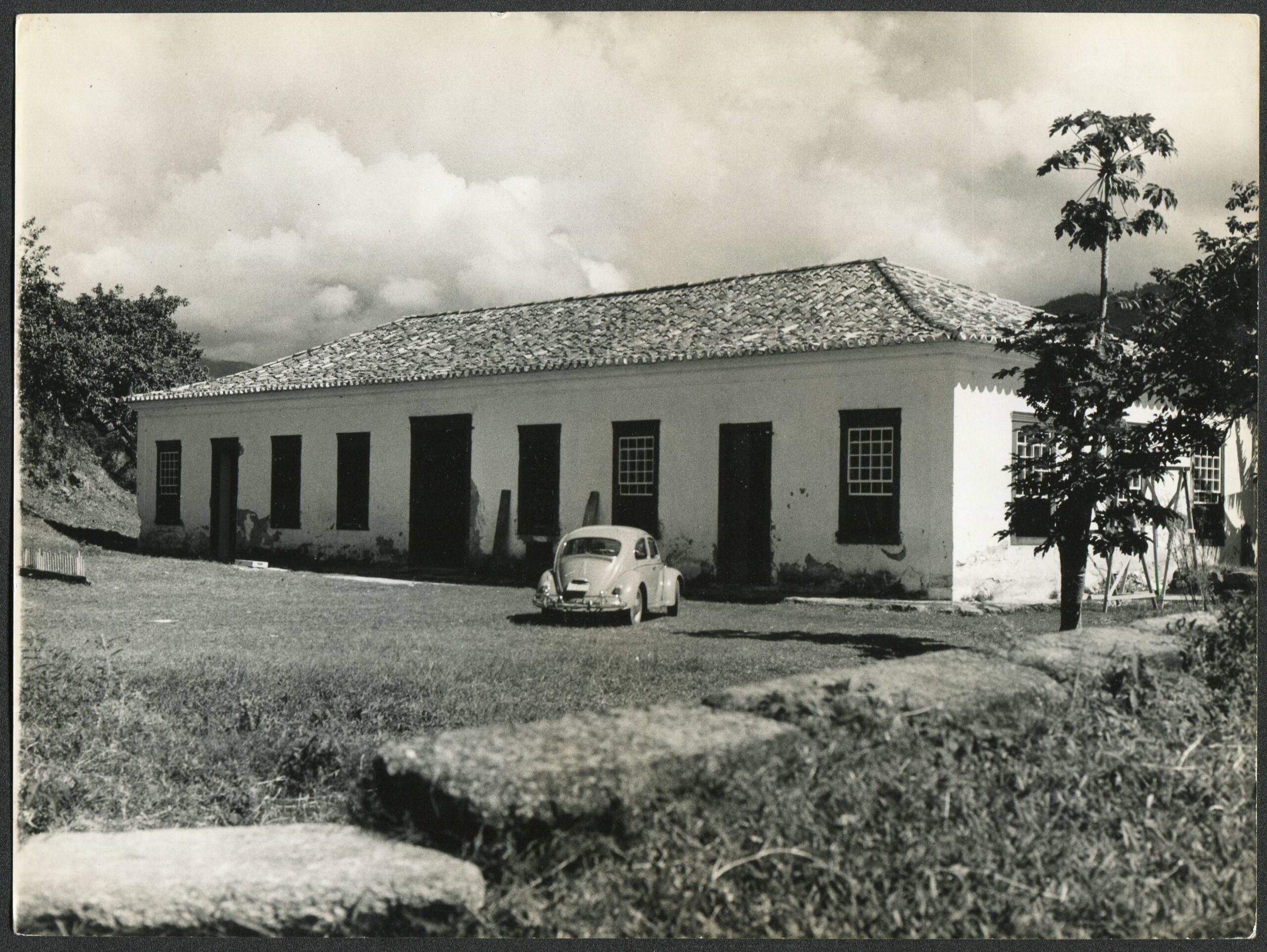 Fotografia histórica em preto e branco, retratando o prédio do museu. No primeiro plano, há um grande gramado e, em frente ao edifício, um carro, um fusca, modelo clássico automobilístico. Ao fundo, grandes nuvens tapam a vista dos morros, caracterizando o dia nublado.