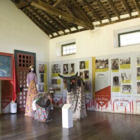 Fotografia da sala expositiva com teto de madeira, sem forro e paredes brancas, com duas janelas pequenas de vidro, dispostas no alto da parede, e piso de madeira escura. Exposição com tema folclórico, com manequins vestidos de roupas tradicionais coloridas e um boi decorado, o Bumba Meu Boi. No fundo, painéis informativos nas paredes, com textos e fotografias que contextualizam os itens expostos.