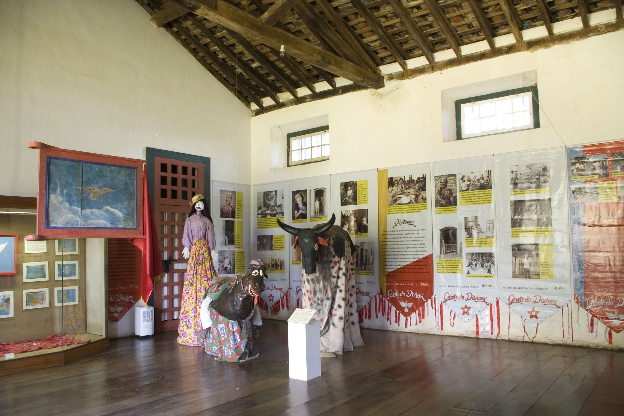 Fotografia da sala expositiva com teto de madeira, sem forro e paredes brancas, com duas janelas pequenas de vidro, dispostas no alto da parede, e piso de madeira escura. Exposição com tema folclórico, com manequins vestidos de roupas tradicionais coloridas e um boi decorado, o Bumba Meu Boi. No fundo, painéis informativos nas paredes, com textos e fotografias que contextualizam os itens expostos.