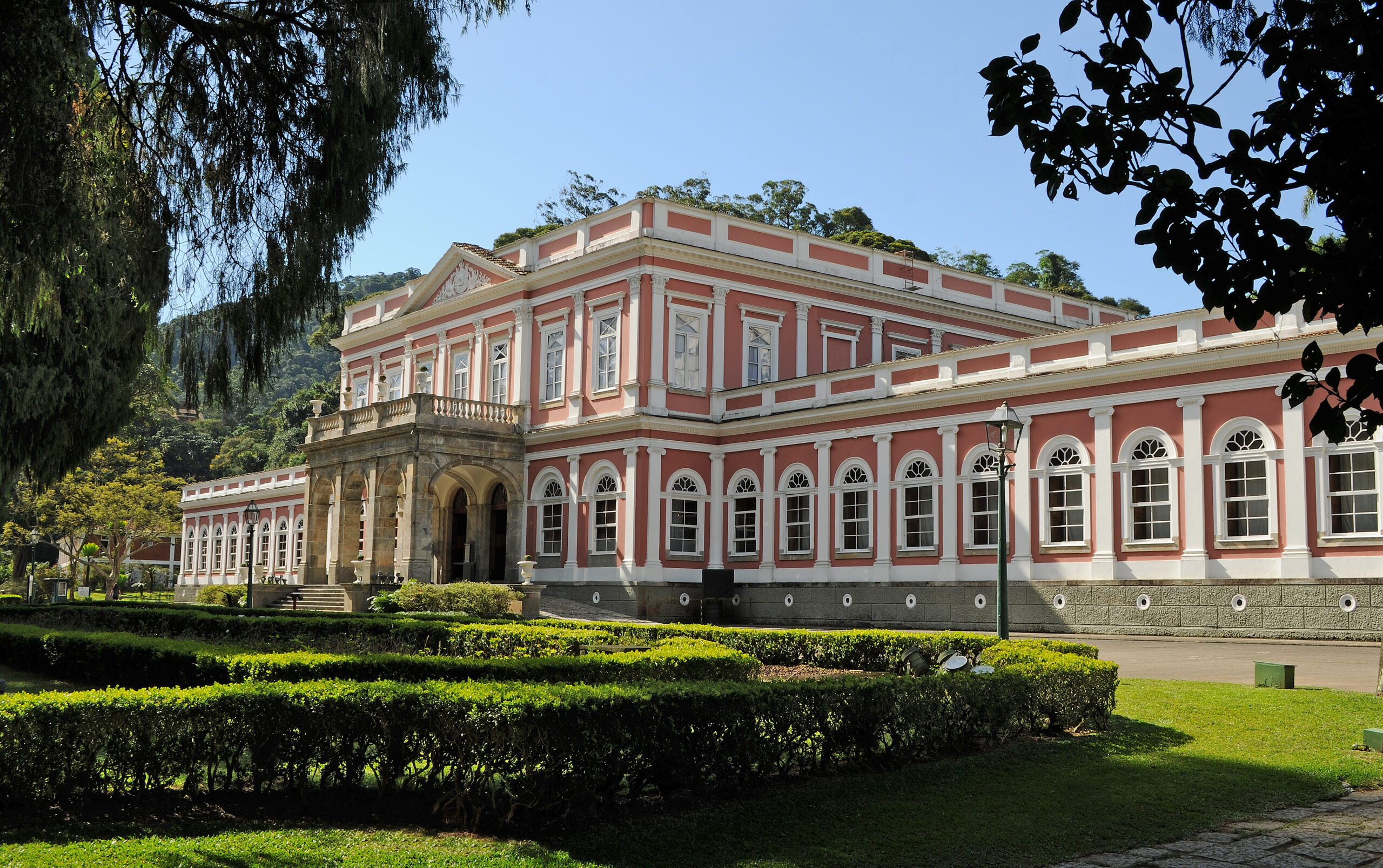 Foto em perspectiva da fachada do Museu Imperial, com destaque para o edifício principal. A construção, de fachada rosa com detalhes brancos, possui dois andares: o térreo apresenta janelas arqueadas, enquanto o andar superior exibe janelas retangulares altas. No centro da fachada, um pórtico com colunas sustenta uma varanda acessada pelo segundo pavimento e demarca a entrada principal. Em frente ao edifício, jardins geométricos compõem o paisagismo do conjunto.