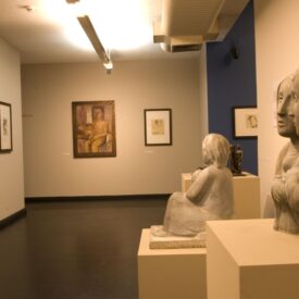 Fotografia do interior do museu. Sala expositiva de parede clara e piso de madeira, com quadros alinhados de vários tamanhos distribuídos nas paredes. Ao fundo, no centro, há um quadro maior com a pintura de uma pessoa sentada. À direita, outro quadro menor, também emoldurado. Em primeiro plano, à direita, duas esculturas de figuras humanas sentadas, posicionadas sobre pedestais quadrados.