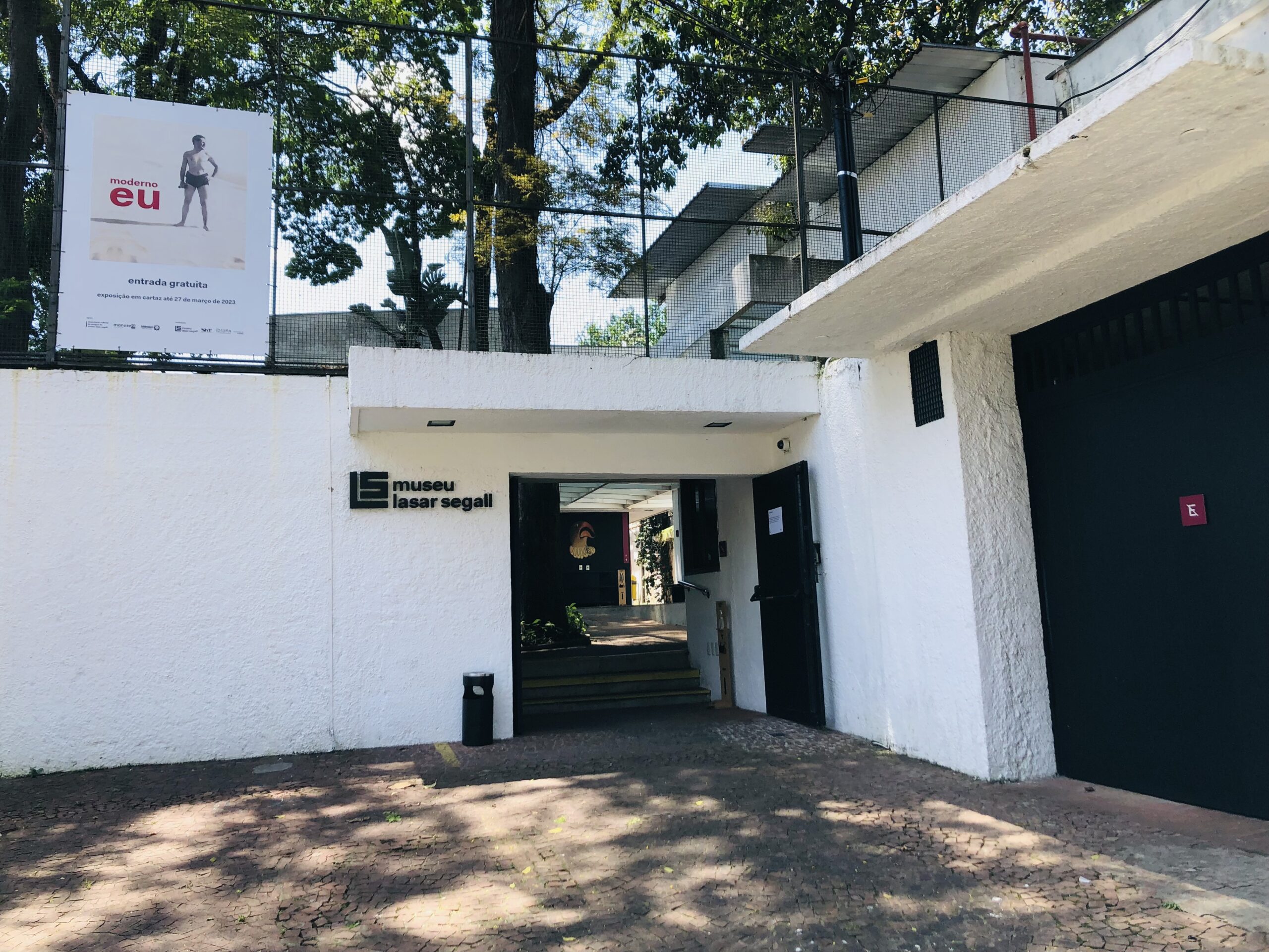 Vista da entrada do museu com um pequeno beiral. O edifício possui uma pintura branca. À esquerda da entrada, há uma placa preta fixada na parede com o nome do Museu. A passagem conduz a um corredor interno, onde é possível ver escadas ao fundo.