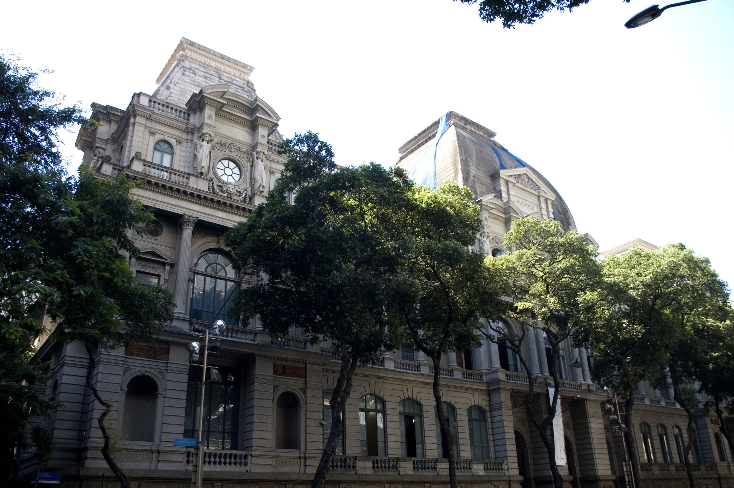 Fotografia da fachada principal do Museu. Seu volume é encimado por duas cúpulas laterais, em menor dimensão, dois terraços e uma cúpula central. Trata-se de edificação imponente, rica em detalhes arquitetônicos, como colunas e janelas arqueadas. As janelas do edifício são de tamanho e formato uniforme, e a fachada em tons de cinza é adornada com ornamentos. O céu é claro e ensolarado, com a luz natural que incide sobre o edifício e as árvores ao redor.