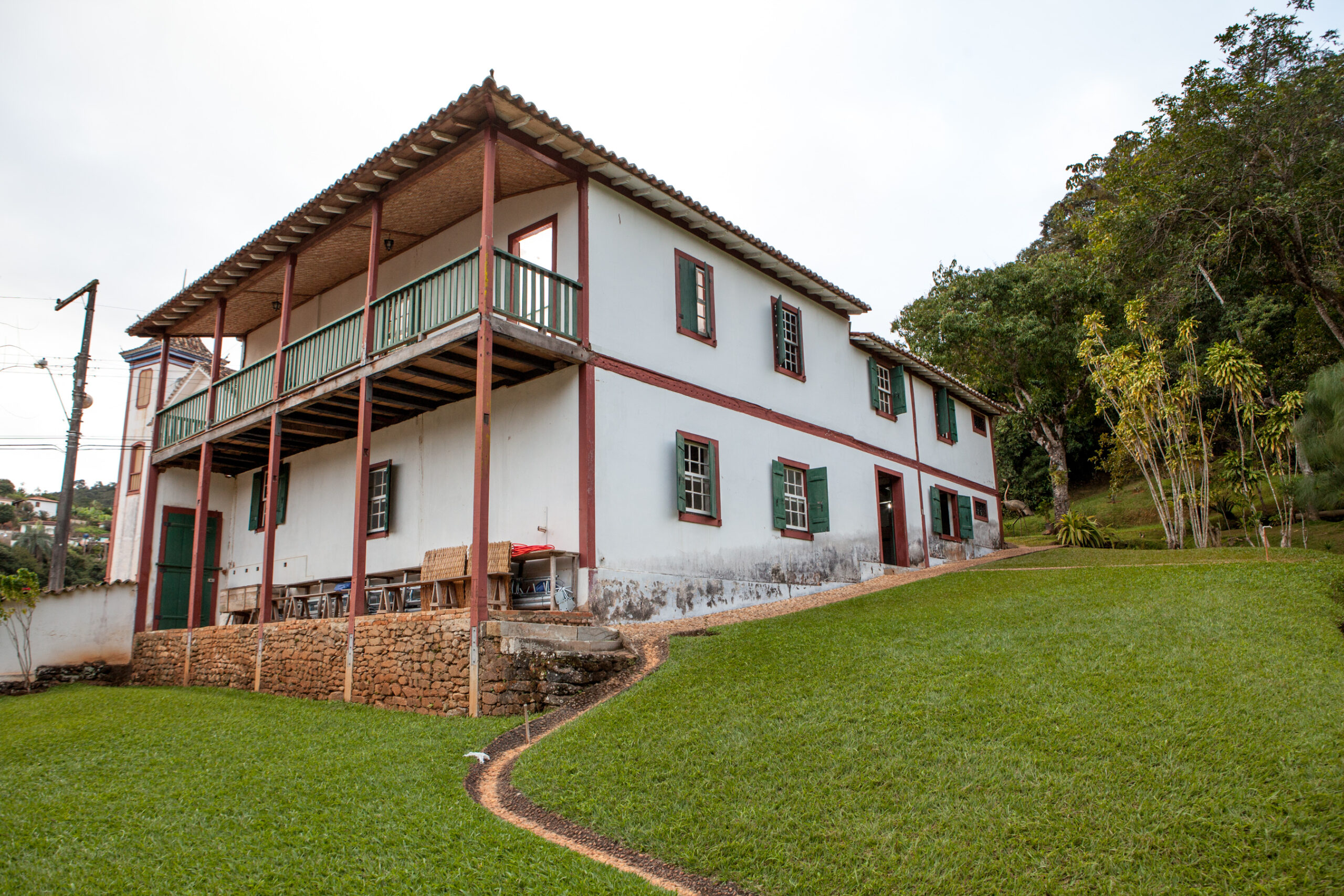 Fotografia do Museu. Edificação de dois andares com paredes brancas e detalhes avermelhados que destacam sua estrutura. As janelas têm pintura verde e molduras avermelhadas. Um grande alpendre orna o segundo andar, sustentado por pilares de madeira no térreo. Em frente à casa, há uma área gramada e, na parte dos fundos, a vegetação mais densa.