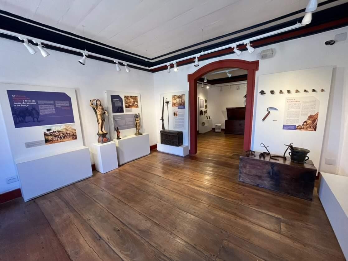 Fotografia da sala expositiva, com paredes brancas e piso de madeira. A iluminação uniforme e bem distribuída destaca as peças sem causar sombras fortes, enfatizando os detalhes históricos expostos em vitrines e painéis. Os objetos e peças históricas incluem esculturas, armas e ferramentas antigas, bem como painéis informativos.