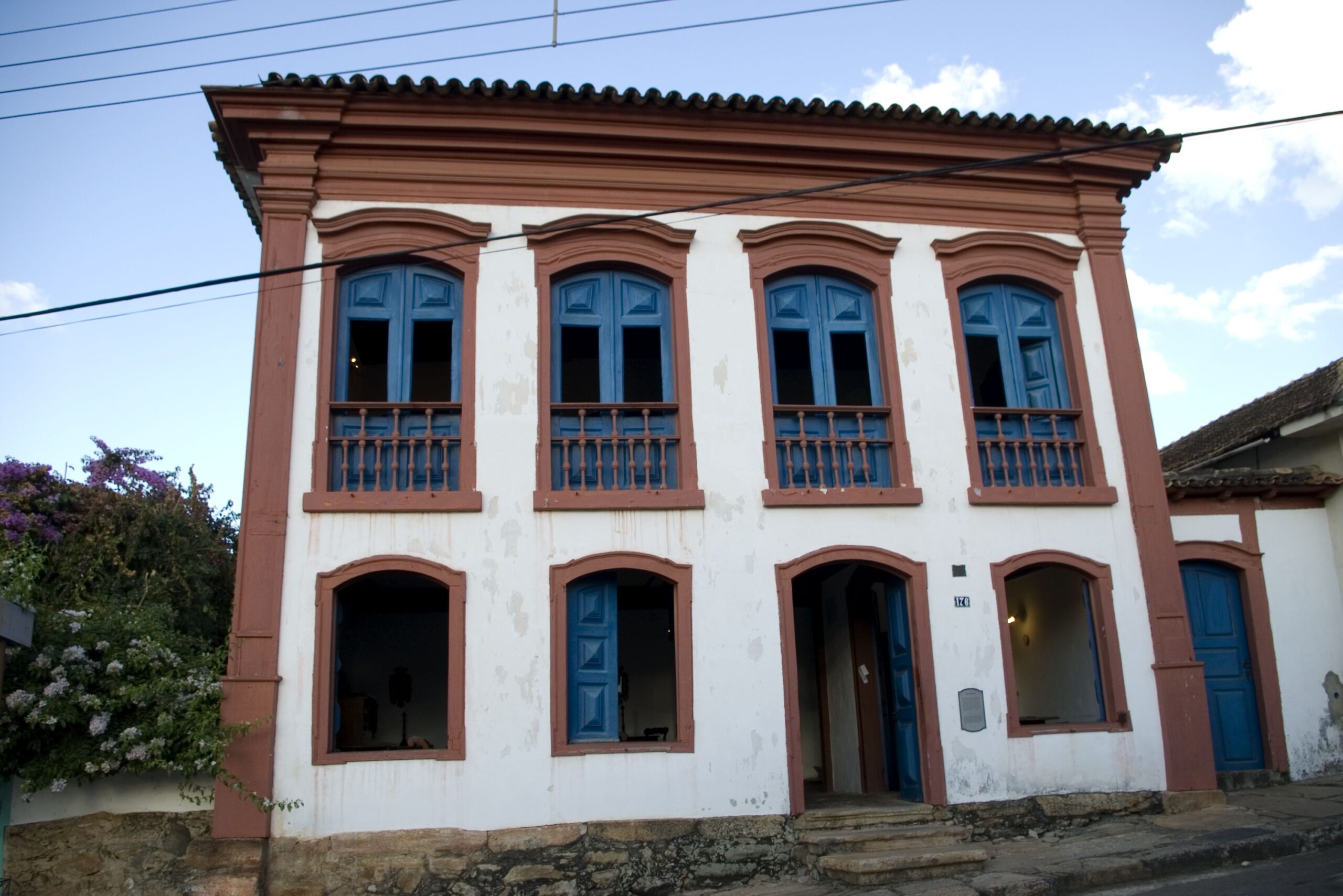Fotografia da fachada do Museu. Fachada de edifício imponente em estilo colonial com pintura na cor branca, com janelas e portas azuis e molduras em vermelho-tijolo. O edifício possui três janelas, uma porta principal, localizadas no térreo, e quatro portas com balcões em balaústre no primeiro pavimento. No acabamento da fachada, nota-se a cimalha desenhada e pintada em vermelho-tijolo.