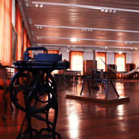 Fotografia da sala expositiva. Sala com piso de madeira polida, com contrastes suaves entre os tons claros das paredes e o tom alaranjado das cortinas. O espaço é amplo e há diversas peças de mobiliário, bem como instrumentos e utensílios, dispostos em diferentes partes da sala. A iluminação é natural, proveniente de janelas.