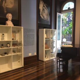 Fotografia da área expositiva. A composição da imagem apresenta uma sala com um piano preto posicionado no centro da cena, próximo à parede direita. Estantes brancas com objetos expostos ocupam a parede esquerda. A parede atrás da estante é de cor azul claro e uma moldura de madeira escura emoldura as paredes e o piso. No canto superior esquerdo, um busto de um homem. A luz natural entra pela janela dupla, localizada à direita do piano.
