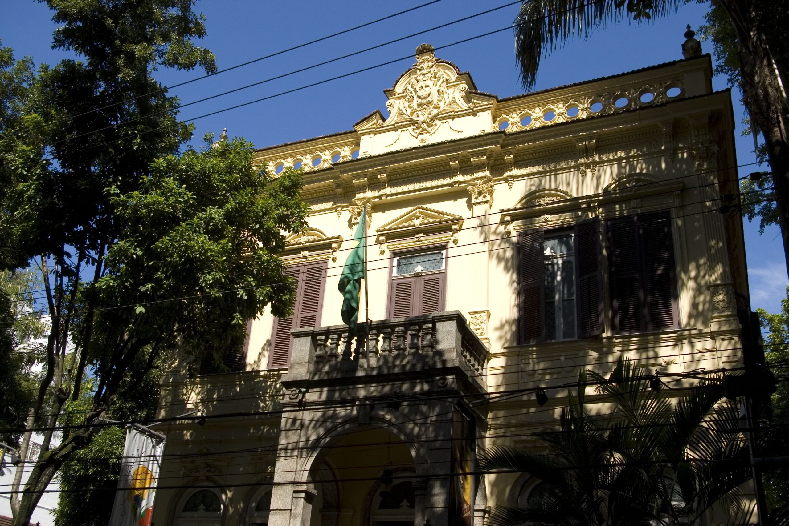 Fotografia do Museu, com foco principal na fachada do edifício, que apresenta janelas com esquadrias marrom-escuras, uma varanda com balaustradas e uma cornija elaborada. As sombras projetadas pelas árvores em volta do edifício criam um contraste entre o edifício e a natureza.